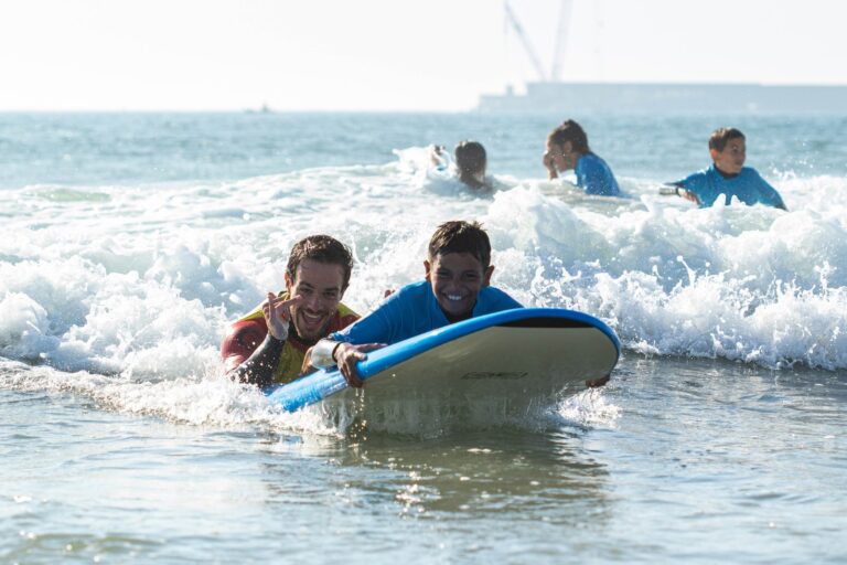 A surf instructor supporting a smiling child on a surfboard during an adaptive surfing session in Porto, Portugal