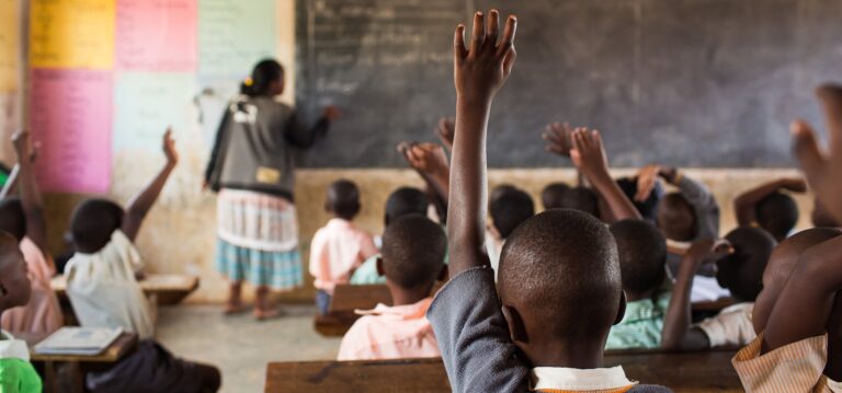 Local teacher at the blackboard with pupils raising their hands in a primary school classroom