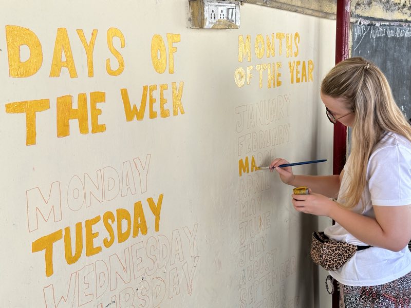 A volunteer painting a days-of-the-week classroom mural